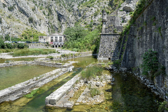 View of stone fortifications and walls of Kotor, with a clear stream and stone structures in the foreground, set against a steep mountain landscape in Kotor, Kotor Municipality, Montenegro.
