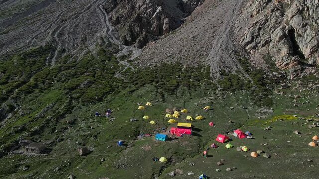 Aerial view of colorful tents scattered on a grassy mountain slope at the base of the massive rocky Diamir face of Nanga Parbat in Diamir, Gilgit Baltistan, Pakistan.