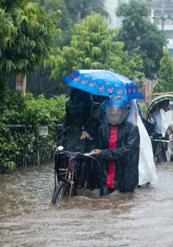 Feni, Chittagong Division, Bangladesh - 08 July 2025: View of people navigating a flooded street during heavy rain, with a man in a raincoat and helmet pushing a bicycle under a blue umbrella.