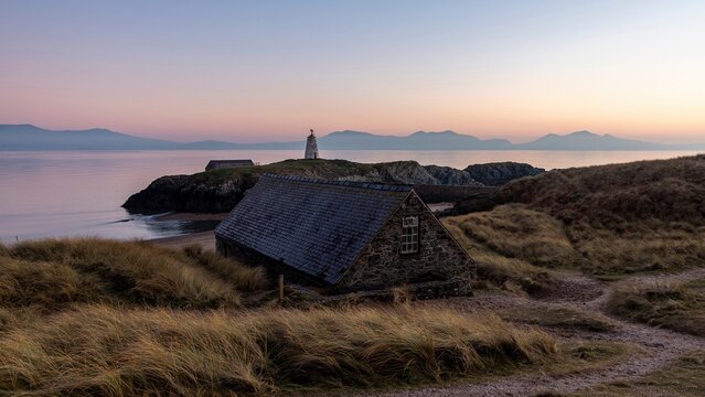 View of Llanddwyn Lighthouse on a rocky island with a stone cottage in the foreground, surrounded by grassy dunes and calm sea at sunset in Caernarfon, Wales, United Kingdom.