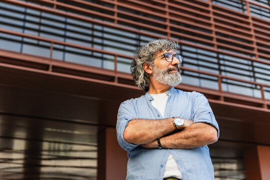 Portrait of senior man standing with crossed arms in front of a modern building in the city, looking pensive to the side
