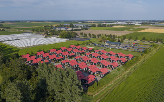 Aerial view of modular housing for migrant and seasonal workers with distinctive red roofs and black walls, situated amidst agricultural fields and greenhouses in Luttelgeest, Flevoland, Netherlands.