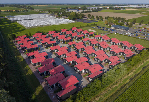 Aerial view of modular housing for migrant and seasonal workers with rows of black cabins and red roofs, surrounded by rural fields and greenhouses in Luttelgeest, Flevoland, Netherlands.