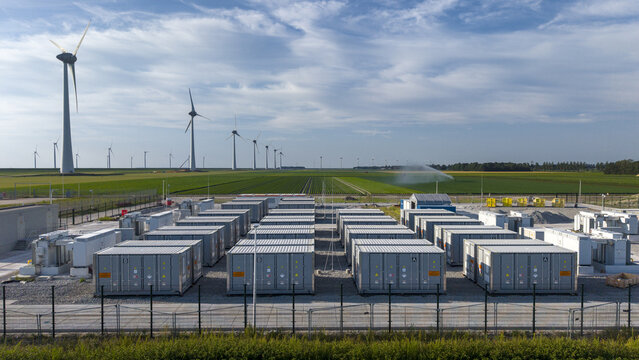 Aerial view of a battery energy storage system with rows of grey containers, wind turbines in the background, and green agricultural fields in Espel, Flevoland, Netherlands.