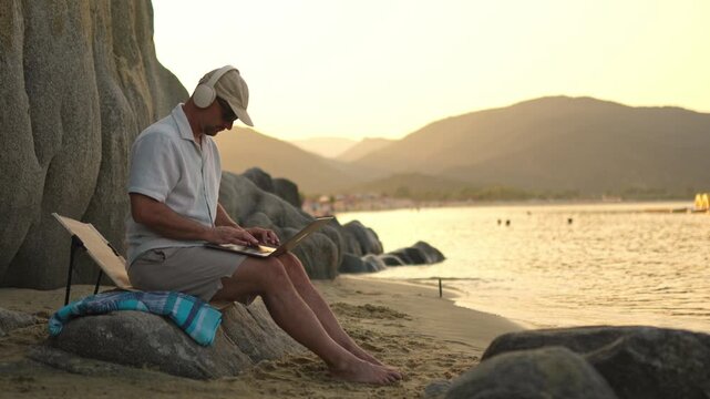 Digital nomad working remotely on a laptop while sitting on rocks by the beautiful sea at sunset