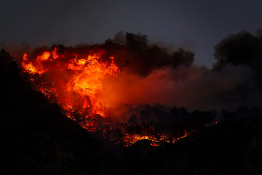 Aerial view of intense forest fires burning through the Susa Valley at night, with bright orange flames and thick smoke rising above the dark landscape in Chianocco, Piedmont, Italy.