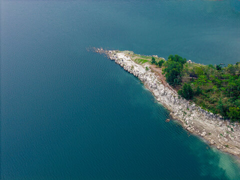 Aerial view of Waduk Wadaslintang featuring a rocky shoreline, lush green vegetation, and deep blue reservoir waters in Kecamatan Wadaslintang, Jawa Tengah, Indonesia.