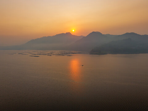 Aerial view of Waduk Wadaslintang at sunset with golden light reflecting on the calm reservoir water, surrounded by misty mountains in Kecamatan Wadaslintang, Jawa Tengah, Indonesia.
