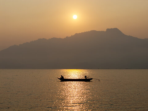 Aerial view of Waduk Wadaslintang with a lone fisherman in a small boat on the calm water during a golden sunset, surrounded by mountains, Kecamatan Wadaslintang, Jawa Tengah, Indonesia.