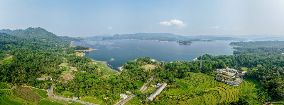 Aerial view of Wadaslintang Reservoir surrounded by lush green hills, terraced rice fields, and a winding road under a clear sky in Wadaslintang, Central Java, Indonesia.