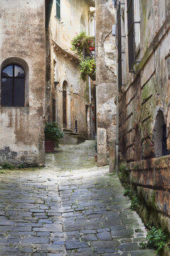 View of a narrow cobblestone alleyway in the historic town of Sorano, featuring weathered stone walls, arched doorways, and potted plants in Sorano, Tuscany, Italy.