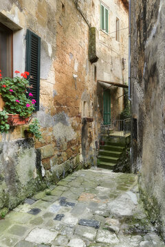 View of a narrow cobblestone alleyway in the historic town, featuring weathered stone walls, green shutters, and a vibrant pot of red geraniums in Sorano, Tuscany, Italy.