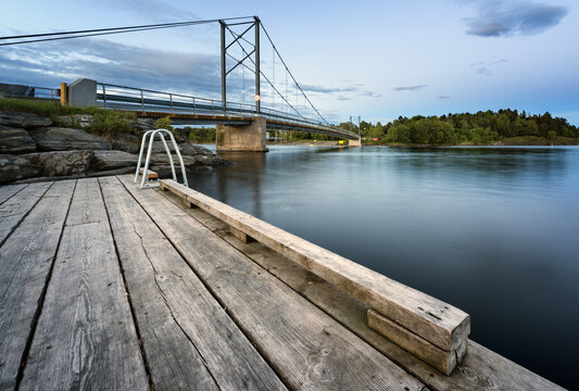 Baerum, Norway - 07 June 2015: View of Kalvoya bridge from a wooden pier with calm water and rocky shoreline under a clear blue sky.