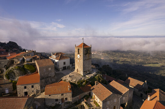 Aerial view of the historic clock tower in the village of Monsanto, surrounded by stone houses with orange tiled roofs, set against clouds in Monsanto, Castelo Branco, Portugal.