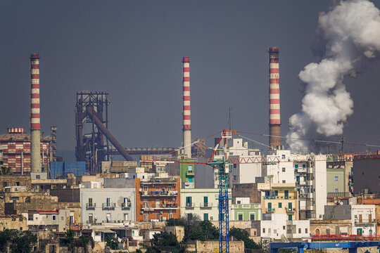 View of the industrial steelworks with tall striped chimneys emitting smoke, rising above the residential buildings and urban landscape of Taranto, Apulia, Italy.