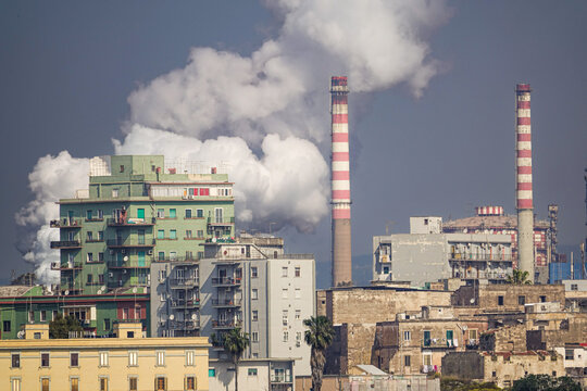 View of the industrial steelworks plant with tall red and white striped smokestacks emitting thick white smoke behind residential apartment buildings in Taranto, Apulia, Italy.