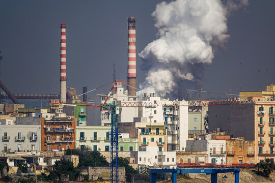 View of industrial chimneys emitting smoke behind residential buildings and a construction crane in Taranto, Apulia, Italy.