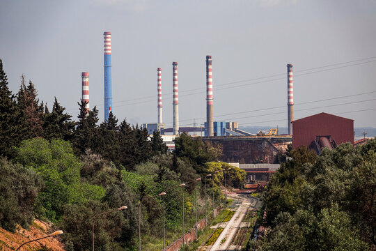 View of the industrial plant with tall striped smokestacks rising above the surrounding trees and railway tracks in Province of Taranto, Apulia, Italy.