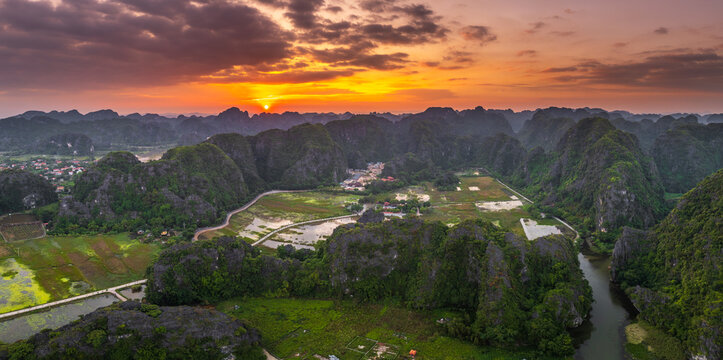 Aerial view of the karst landscape of Tam Coc Bich Dong with winding river, green rice paddies, and limestone mountains under a vibrant sunset sky in Nam Hoa Lu, Ninh Binh, Vietnam.