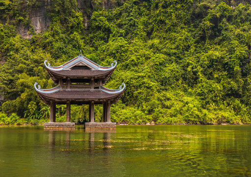 View of the traditional Thuy Dinh pavilion floating on the calm waters of Trang An, surrounded by lush green limestone mountains in Hoa Lu, Ninh Binh, Vietnam.
