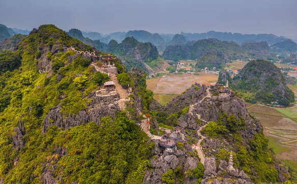 Aerial view of Mua cave with its iconic stone staircase winding up the limestone karst mountain, surrounded by lush greenery and pagodas in Nam Hoa Lu, Ninh Binh, Vietnam.