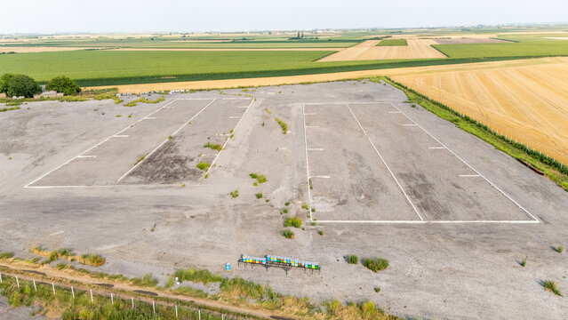 Aerial view of a large gravel lot with white painted rectangular plots surrounded by vast agricultural fields and a row of colorful beehives in Serbia.