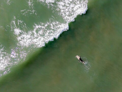 Aerial view of a lone surfer paddling on a surfboard in the green ocean water near a breaking wave in Bigbury-on-Sea, England, United Kingdom.