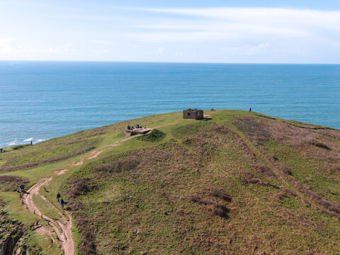 Aerial view of Burgh Island featuring a historic stone bunker atop a grassy hill overlooking the sea with people walking along the coastal path in Bigbury-on-Sea, England, United Kingdom.
