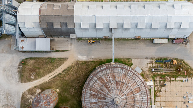 Aerial view of an industrial grain storage facility with large metal silos, a long warehouse, and a circular structure, surrounded by gravel paths in Sremska Mitrovica, Vojvodina, Serbia.