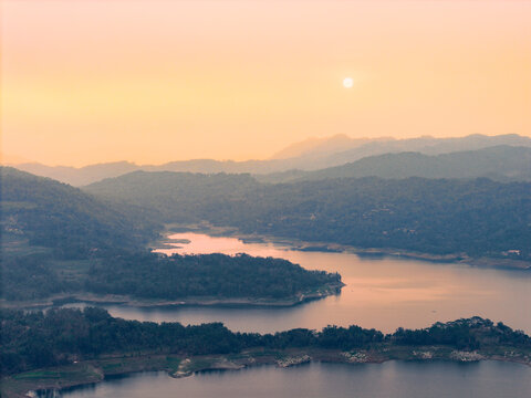Aerial view of Wadaslintang Reservoir at sunset with calm water reflecting the golden light, surrounded by lush green hills in Wadaslintang, Central Java, Indonesia.