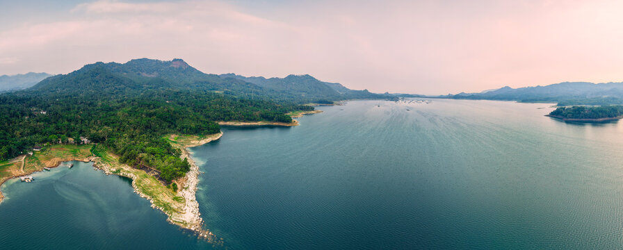 Aerial view of Wadaslintang Reservoir with lush green hills, calm blue water, and rocky shoreline in Wadaslintang, Central Java, Indonesia.