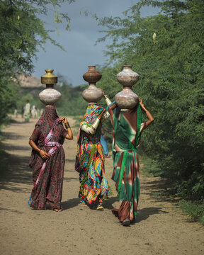 View of three women in traditional colorful clothing carrying water pots on their heads while walking along a dirt path in Nagarparkar, Sindh, Pakistan.