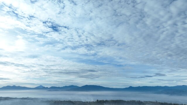 Aerial view of the misty mountainous landscape of Lembang with distant peaks rising above a thick blanket of fog under a vast textured sky in Lembang, West Java, Indonesia.