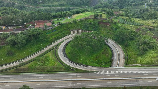 Aerial view of the curved Parungkuda toll road interchange surrounded by lush green hills and vegetation in Parungkuda, West Java, Indonesia.