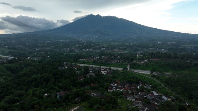 Aerial view of Parungkuda nestled at the base of a mountain, featuring lush vegetation, scattered houses, and a winding road under a cloudy sky in Parungkuda, West Java, Indonesia.