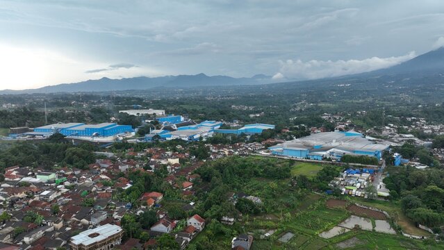 Aerial view of the industrial facility with blue-roofed buildings situated amidst lush greenery, residential houses, and distant mountains in Parungkuda, West Java, Indonesia.