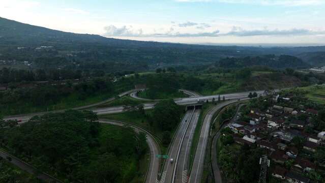 Aerial view of the highway interchange and tunnel entrance surrounded by lush green hills and residential houses in Parungkuda, West Java, Indonesia.
