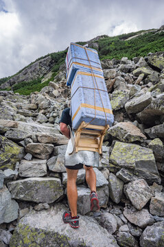 Vysoke Tatry, Slovakia - 22 July 2016: View of a mountain porter carrying a heavy load in a wooden frame up a rocky mountain trail in the High Tatras, with rugged terrain and cloudy skies.