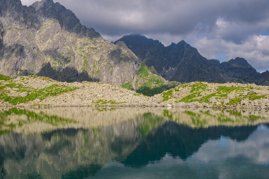 View of Litvorove pleso reflecting the dramatic high peaks of the High Tatras mountains with rocky shores and green slopes under a cloudy sky in Tatranska Javorina, Presovsky kraj, Slovakia.