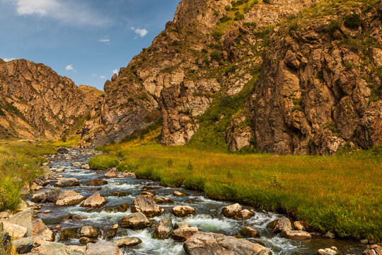 View of a mountain river flowing through a rocky valley with green grassy banks under a blue sky in Sarybulak, Osh Region, Kyrgyzstan.