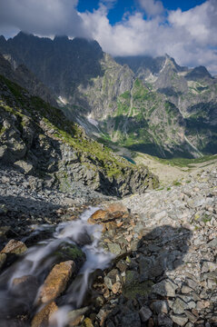 View of Litvorova dolina featuring a cascading mountain stream flowing over rocky terrain with dramatic peaks and clouds in the background in Tatranska Javorina, Presovsky kraj, Slovakia.