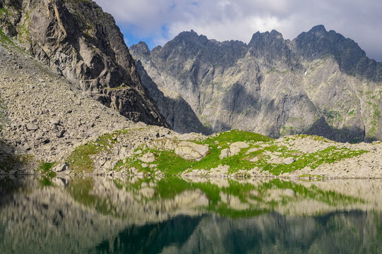View of Litvorove pleso reflecting the rugged peaks of the High Tatras mountains with rocky shores and green grass in Tatranska Javorina, Presovsky kraj, Slovakia.