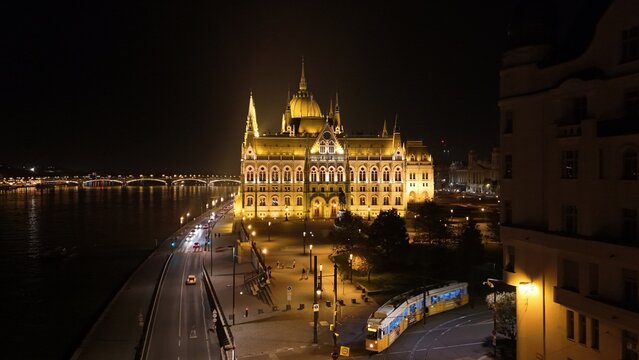 Aerial view of the illuminated Hungarian Parliament Building at night along the Danube River with a tram passing by in Budapest, Hungary.