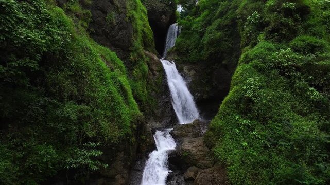 Aerial view of Curug Jagapati waterfall cascading down lush green cliffs into a natural pool in Cisompet, West Java, Indonesia.