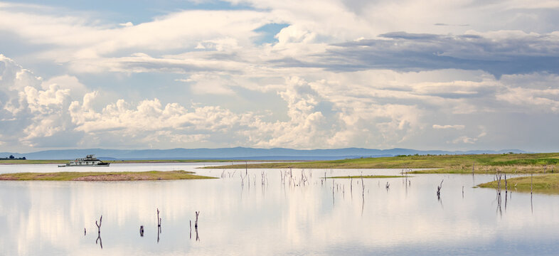 View of a boat on Lake Kariba surrounded by dead trees in the water, grassy banks, and a cloudy sky in Kariba, Mashonaland West Prov, Zimbabwe.