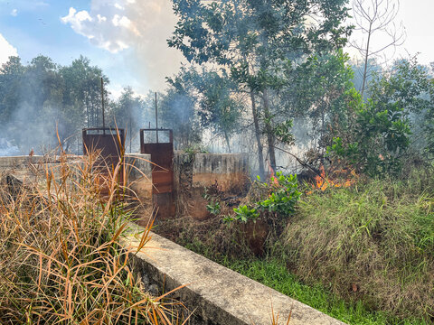 Liang Anggang, Indonesia - 15 July 2026: View of a concrete irrigation structure surrounded by trees and vegetation affected by a forest fire with smoke and flames visible in the background.