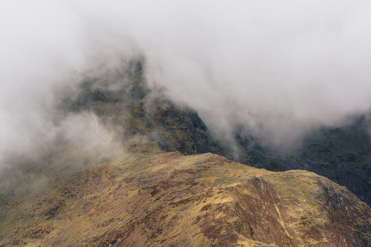 View of Y Lliwedd mountain ridge partially obscured by thick low-hanging clouds and mist, showing rugged rocky terrain and sparse vegetation in Caernarfon, Wales, United Kingdom.
