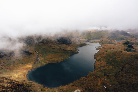 View of the dark waters of Llyn Llydaw nestled among the rugged, mist-covered peaks of the Snowdonia mountain range in Caernarfon, Wales, United Kingdom.