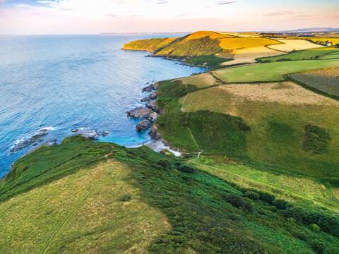 Aerial view of Lansallos Beach with rugged coastal cliffs, lush green fields, and blue sea under golden hour light in Looe, Cornwall, United Kingdom.