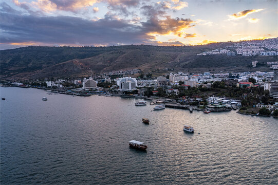 Aerial view of Tiberias on the shore of the Sea of Galilee with boats on the water, hotels, and urban buildings under a sunset sky in Tiberias, North District, Israel.